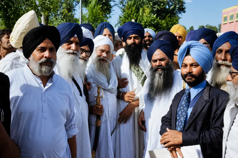 A large group of Sikh men gathered outdoors in bright sunlight, one carrying a kirpan.