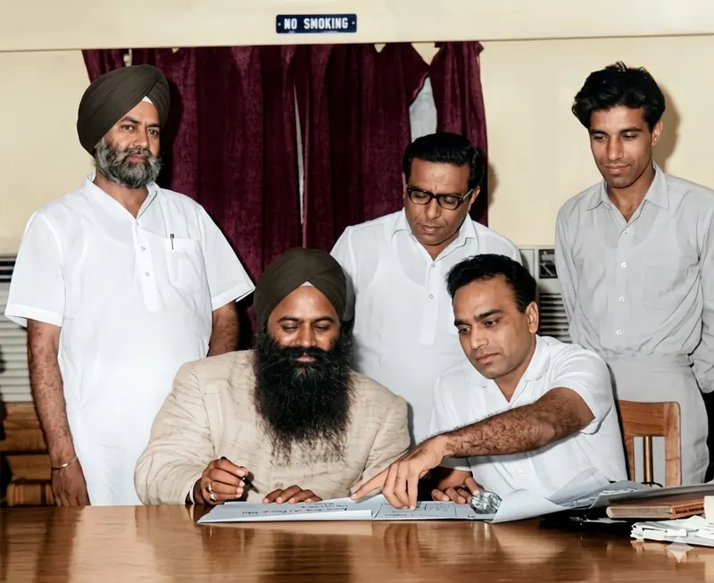 Seated at a desk signing a document, a DDA official beside him pointing to the page, three men standing behind.